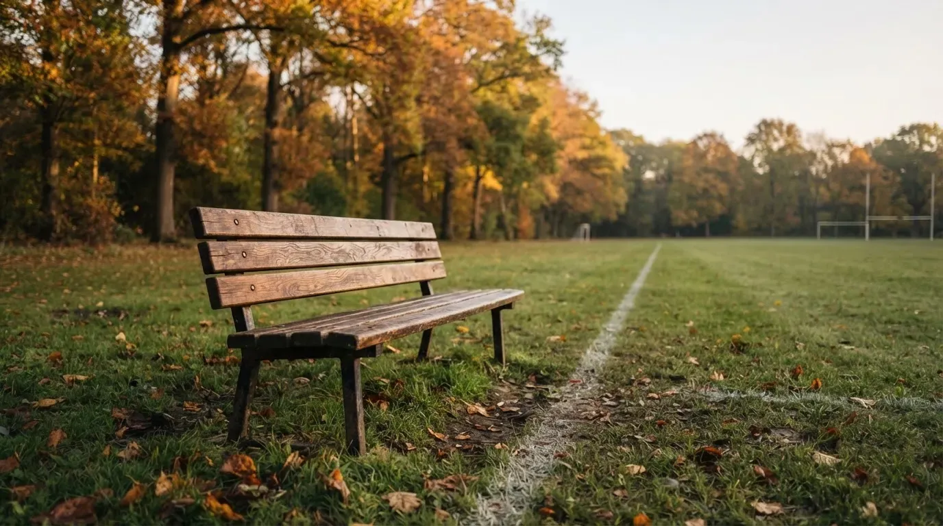 Eine hölzerne Bank steht an der Seitenlinie eines leeren Sportplatzes, dahinter Bäume im Herbstlicht.