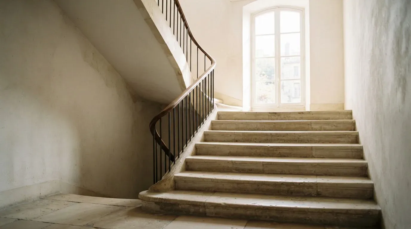 Ein Treppenhaus aus hellem Stein führt in einem Altbau nach oben, Licht fällt durch ein hohes Fenster.